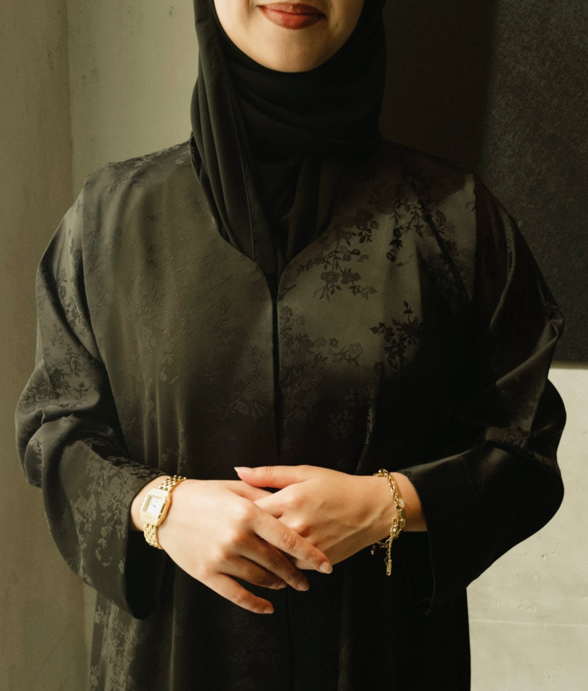 A woman wearing a black long-sleeve abaya with a subtle tonal floral jacquard pattern and a matching black hijab, accessorized with gold bracelets and a watch.